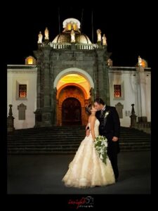 A bride and groom kiss in front of a cathedral at night