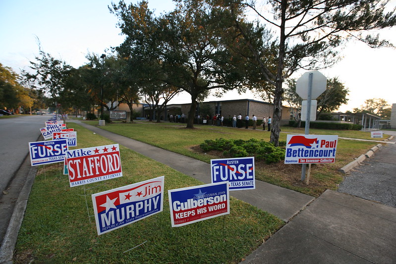 Various campaign signs are propped into the ground around a sidewalk.