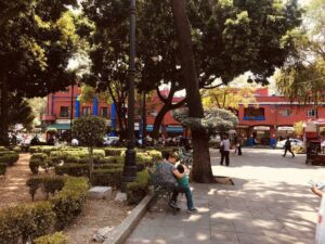 Photo of a person with a child sitting on a bench in a city with trees and bushes nearby. There is a red building in the background.