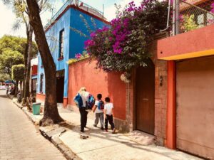 A photo of an adult and two kids with backpacks walking down a sidewalk, next to a bright red building and bright blue building.