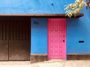 A photo of a hot pink door on a bright blue wall, with house number "105" in black on the wall.