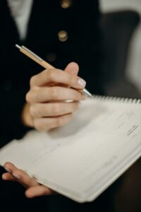 A hand poised to write, holding a light orange pen over a spiral-bound notebook.