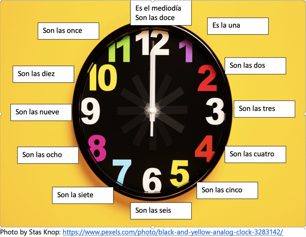 Photo of a black clock. At each number there is a label for how to tell the time in Spanish. Starting with 12 and going clockwise, these read: es el mediodía. Son las doce. Es la una. Son las dos. Son las tres. Son las cuatro. Son las cinco. Son las seis. Son las siete. Son las ocho. Son las nueve. Son las diez. Son las once.