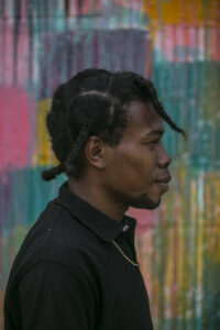A dark-skinned man with braids in his hair wearing a black polo shirt, in profile