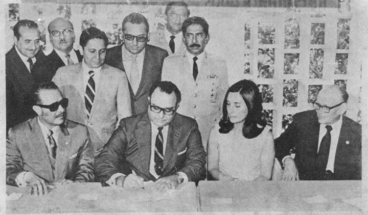 A black-and-white photograph of a man in a suit signing a document, surrounded by others .