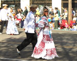 Two people walking down the street, one in a white coat and black pants, and the other in a long white and red patterned dress