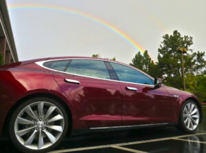 a red Tesla, in a parking space with a rainbow in the gray sky behind the car.