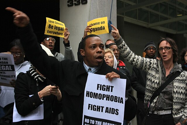 A diverse group of protesters demonstrate against Uganda's Anti-Homosexuality Bill outside a building. At the forefront, a man holds a sign that reads "FIGHT HOMOPHOBIA, FIGHT AIDS – NO TO THE UGANDA..." (partially obscured).