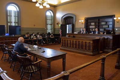 A man sits at a wooden table facing a panel of five people sitting at a wooden desk