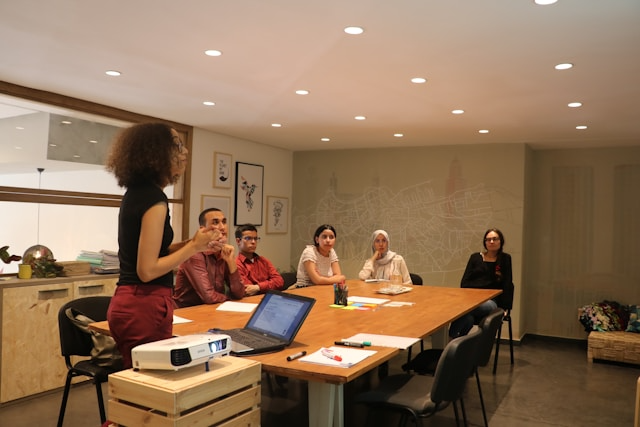 People sitting around a table, looking at a screen, while one woman stands and talks
