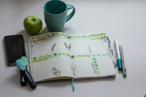Green mug, apple, phone, pens, and an open planner