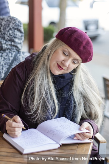 A woman sits at a table and looks down at an open notebook with a pen in her hand