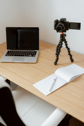 A table holding a laptop computer, a camera on a tripod, and a notebook