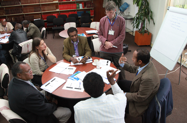Five people sit around a table talking while one man stands and listens