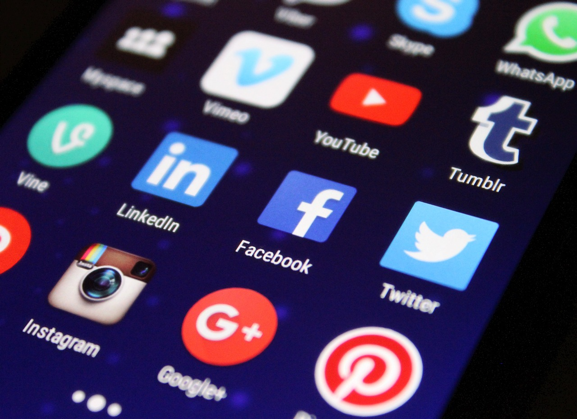 Close-up of a smartphone screen displaying a grid of social media app icons, including Vine, LinkedIn, Instagram, Google+, Pinterest, Vimeo, Facebook, Twitter, YouTube, Tumblr, WhatsApp, and Skype, against a dark background with small blue lights.