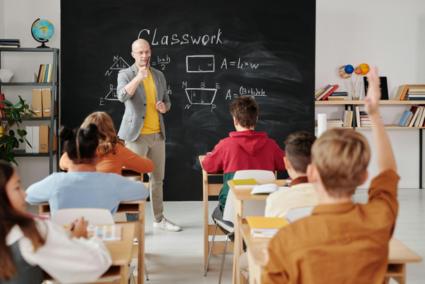 A teacher stands near the blackboard and calls on a student who has their hand raised