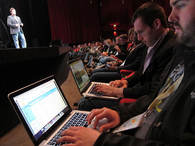 Audience members look at their laptop computers while a person speaks onstage